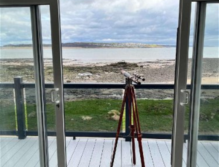 View from the deck of a static caravan at Trecco Bay Holiday Park | A telescope set up on a wooden deck, looking out through large glass doors towards a rocky coastline and calm sea under a cloudy sky.