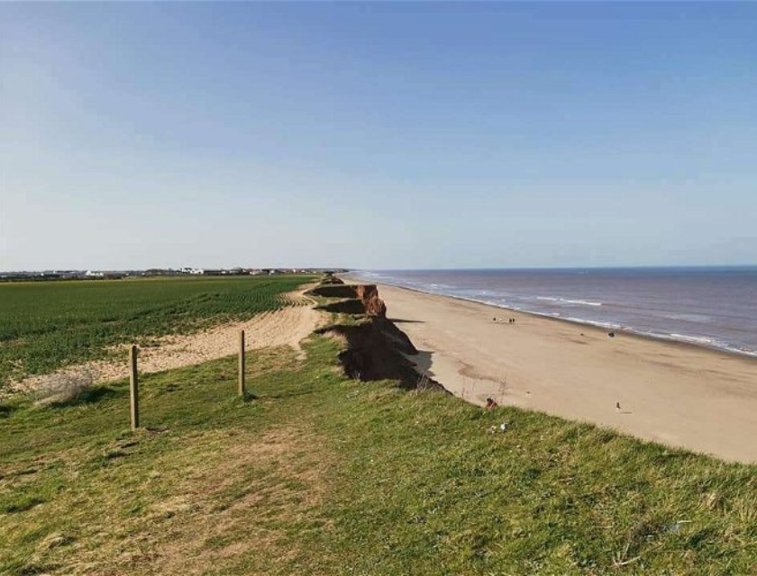 Beach in Withernsea with sand and sea visible on the right, and shore & grass on the left. In the background, the sky is blue with no clouds. 