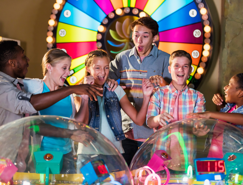 Teens kids enjoying themselves at an arcade. There is a colourful digital spinning wheel behind them. There are 6 people in total, 3 boys and 3 girls.