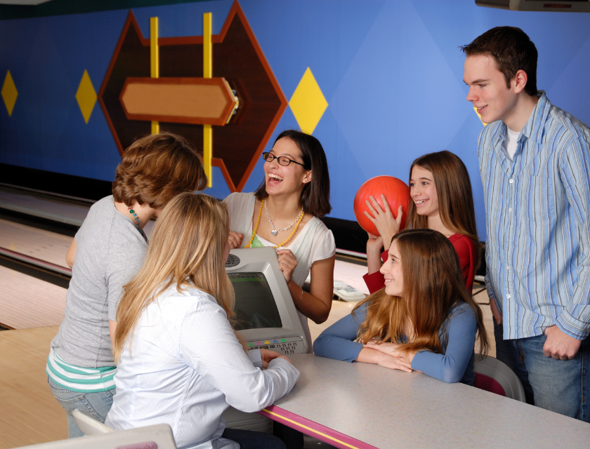 Teens around a screen at a bowling alley. There are 6 people in total, 4 girls and 2 boys, one girl is holding a red bowling ball.