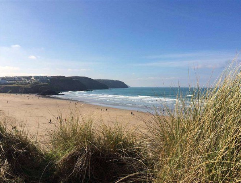 The perfect base for finding cycling trails in Cornwall. A beach from behind some tall grass with a bright blue sky in the background.