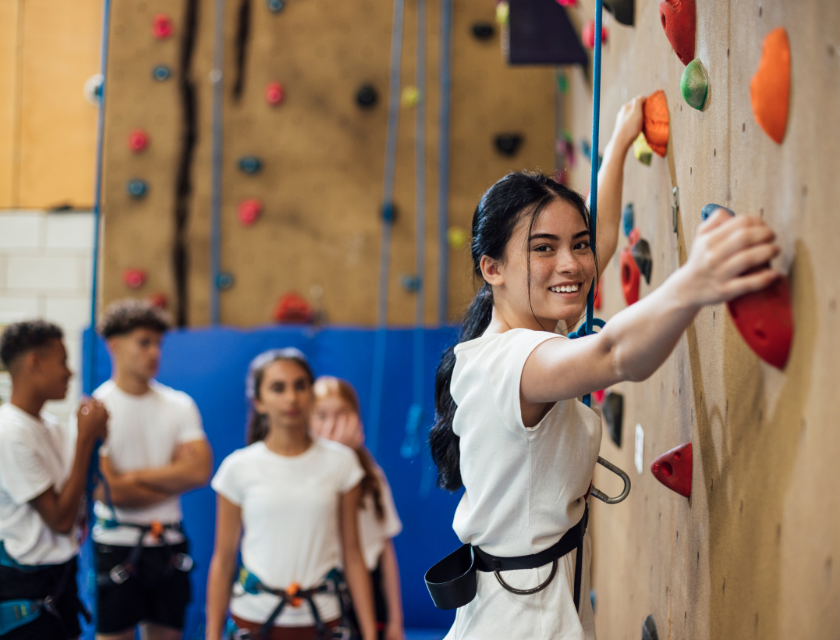 A group of people climbing on a climbing wall. A girl is looking directly into the camera and smiling.