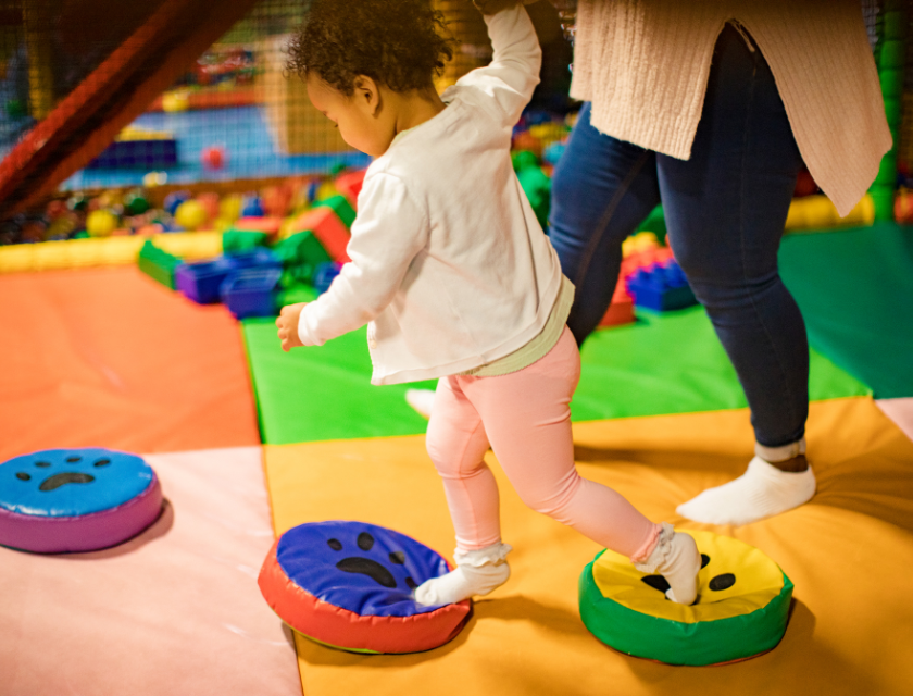 Toddler holding an adults hand while the toddler steps on toys in a soft play area.