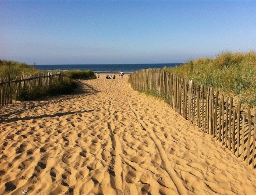 Sandy pathway leading to the sea at Golden Sands Holiday Park near Lincolnshire. The bright sky is blue and there is a wooden fence at either side of the sandy pathway. 