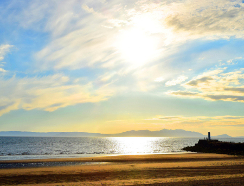 A landscape of the beach in Ayr Scotland with silhouette of the harbour wall, shimmering sea, distant isle of Arran, the setting sun and sky of pale yellow and blue with the use of colour saturation.