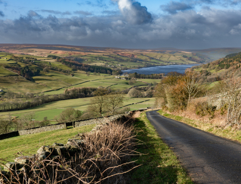 Concrete road going downhill in Nidderdale, Yorkshire . There is shrubbery and grass on both sides of the road. The sky is filled with grey clouds.