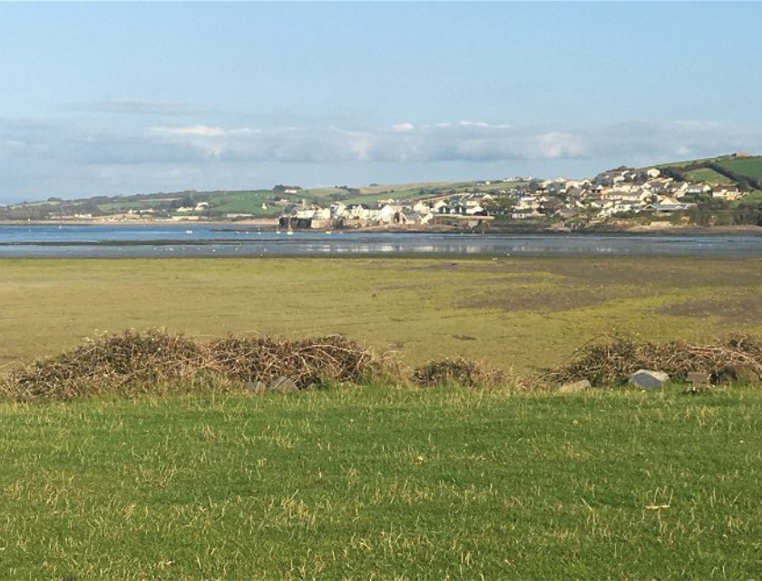 A scenic view from Surf Bay Holiday Park near Woolacombe Beach, showing a grassy field leading to a calm body of water with a coastal town and rolling hills in the background under a clear sky.