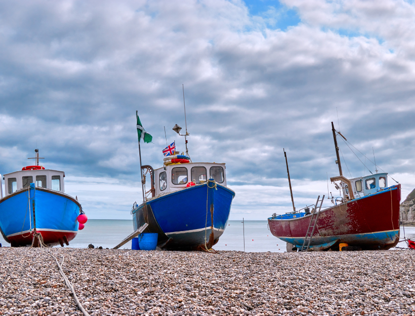 Three colorful fishing boats moored on a pebble beach, under a cloudy sky, with the ocean visible in the background