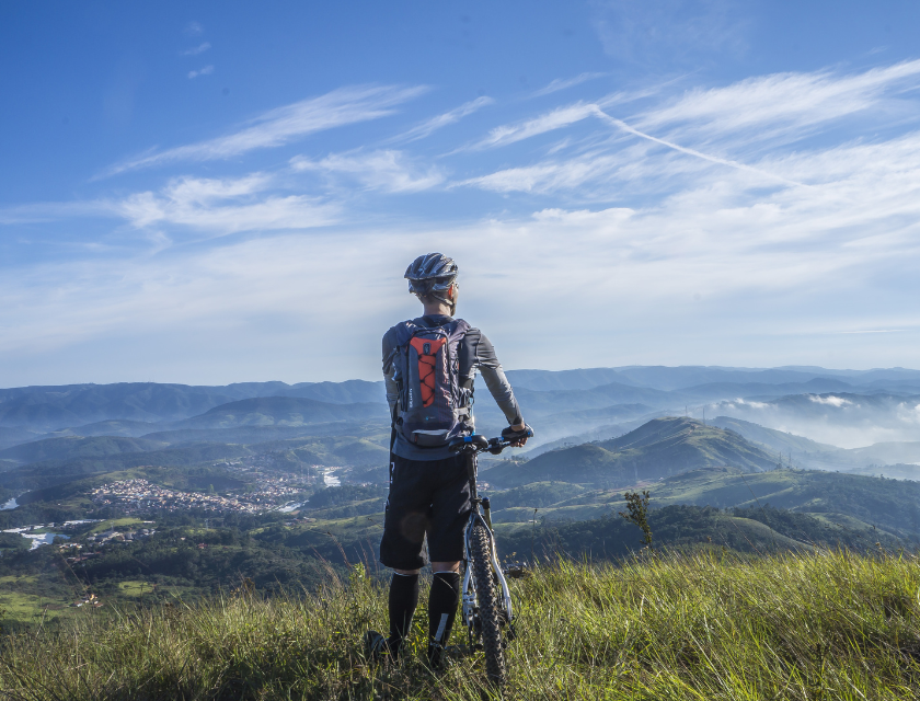 A cyclist wearing a helmet and backpack stands on a grassy hilltop with their bike, looking out over a vast landscape of rolling hills and distant mountains under a bright blue sky, capturing the sense of adventure and panoramic views that come with cycling.