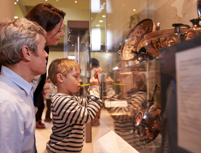 Family observing and interacting with historical artifacts in a museum display case, with a young boy focused on the exhibit. Example of a rainy day thing to do in Cornwall for families. 