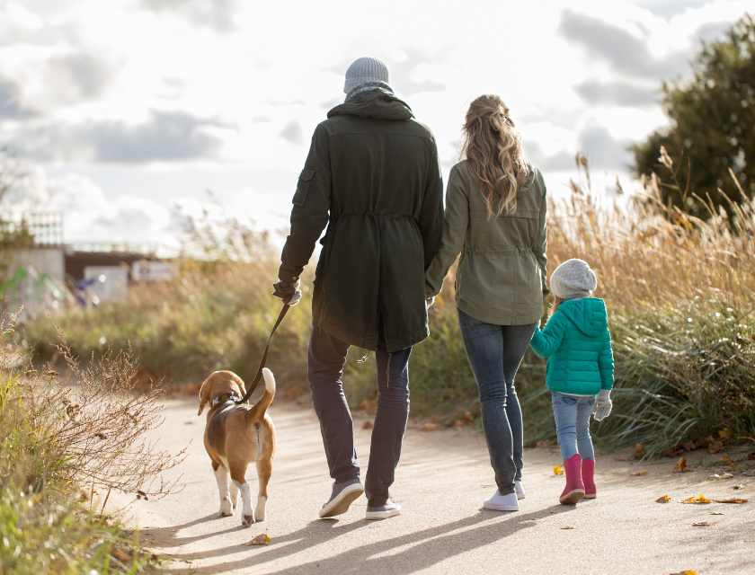 Man, woman and child dressed in autumnal clothing taking a walk through a sandy pathway with shrubs at either side. The man has a leashed dog on his left arm.