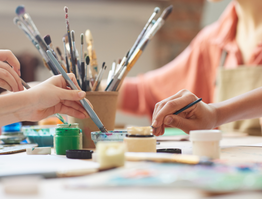 Close up of a paint pot in an arts & crafts class. Several hands holding paintbrushes & dipping into the paint pot.