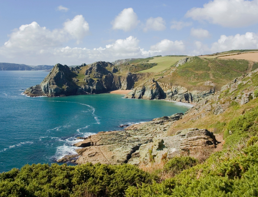 Rocky coastline with a small secluded beach and cliffs, surrounded by green fields and shrubs, under a partly cloudy sky in Devon, England.