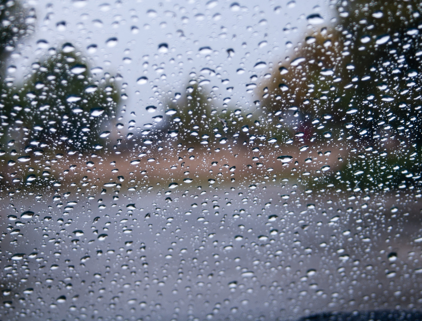 Close-up view of raindrops on a window, with a blurred outdoor scene of trees and a road in the background.