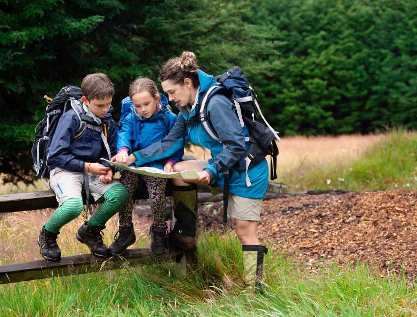 Family of three hikers in outdoor gear looking at a map, standing on a trail with lush green trees in the background.