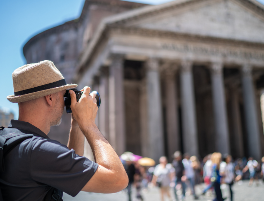 A man wearing a hat and backpack takes a photograph of an ancient building with tall columns, likely a historic landmark, as tourists walk by in the background