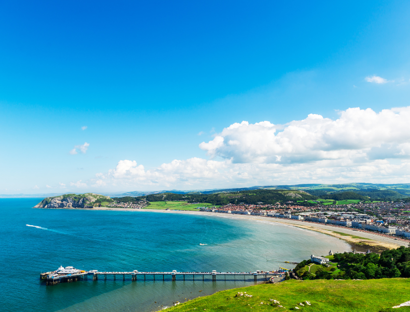 North Wales is a great destination for beach lovers and coastal walks. Image shows an aerial view of a beautiful bay with a long pier extending into the calm blue sea, backed by rolling green hills and a charming coastal town. 