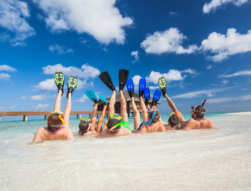 A group of snorkelers lying on the edge of the water with their colorful flippers raised in the air, enjoying a bright, sunny day with a clear blue sky.