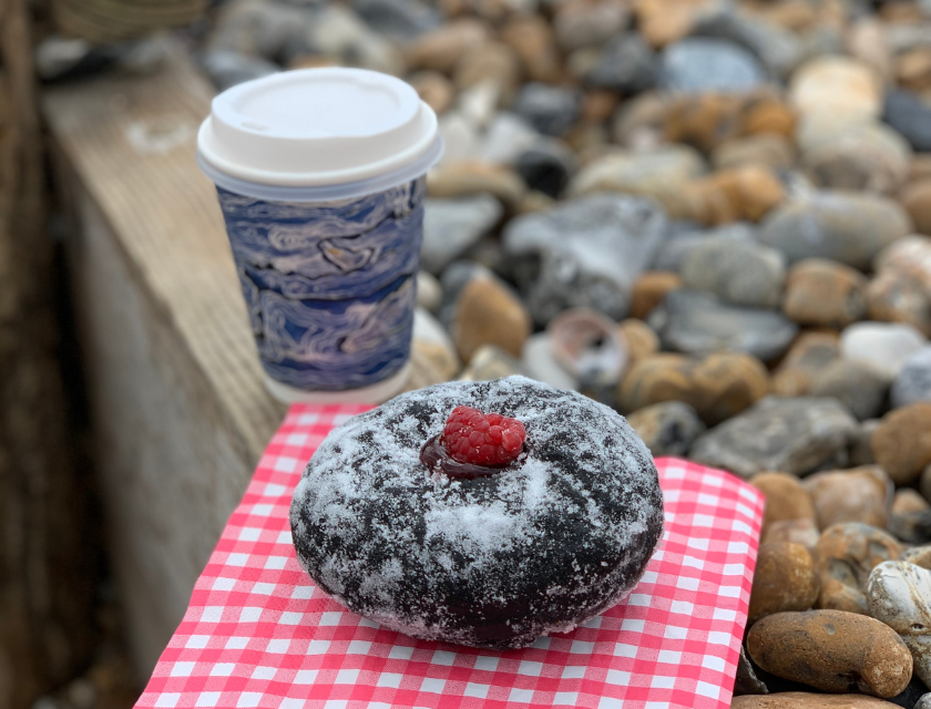 Close-up of a powdered chocolate pastry topped with a fresh raspberry on a red and white checkered cloth. A to-go coffee cup sits in the background, with smooth stones scattered on the surface.