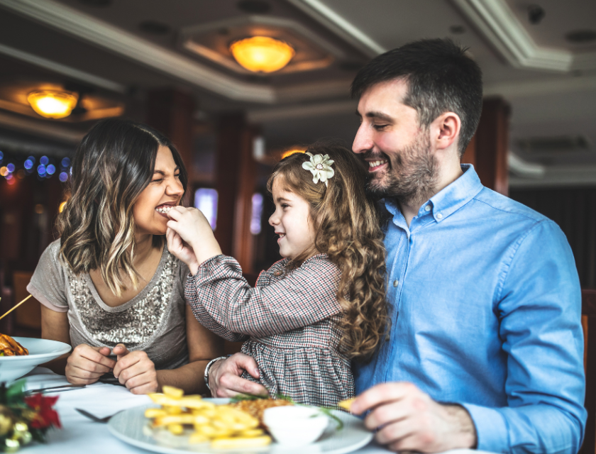 A happy family dining at a restaurant. The young girl feeds her smiling mother while sitting on her father's lap. Everyone is smiling.