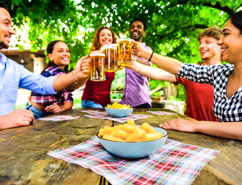 A group of friends sitting outdoors at a wooden table, raising glasses of beer in a toast, with bowls of snacks in the foreground. 