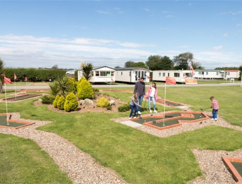 Facilities at Withernsea Sands Holiday Park include crazy golf. Family of 3 with a young boy playing mini golf with a static caravan park in the background.