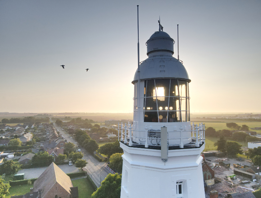 Aerial shot of the Withernsea Lighthouse Museum on a sunny day as the sun sets. There are two birds in the background and homes are visible below.