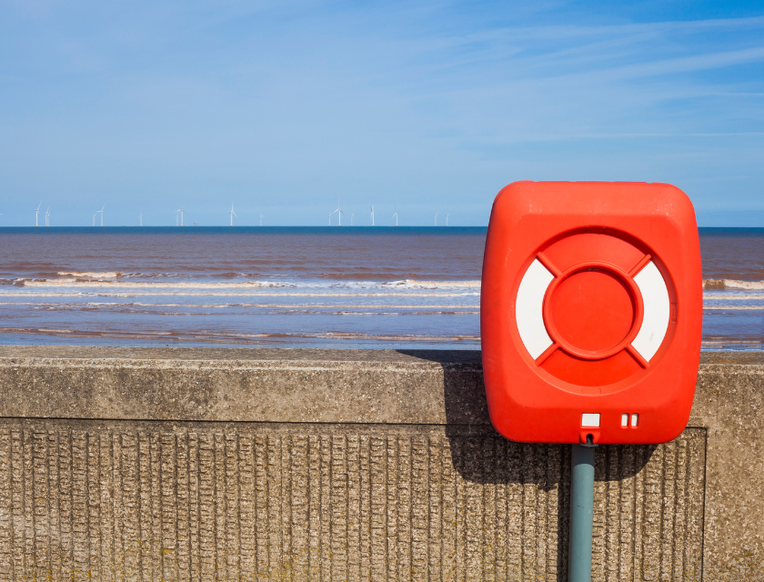 A lifebuoy in its square housing, on a concrete sea wall overlooking the ocean. In the background, the sea stretches to the horizon, with wind turbines visible in the distance under a clear blue sky.