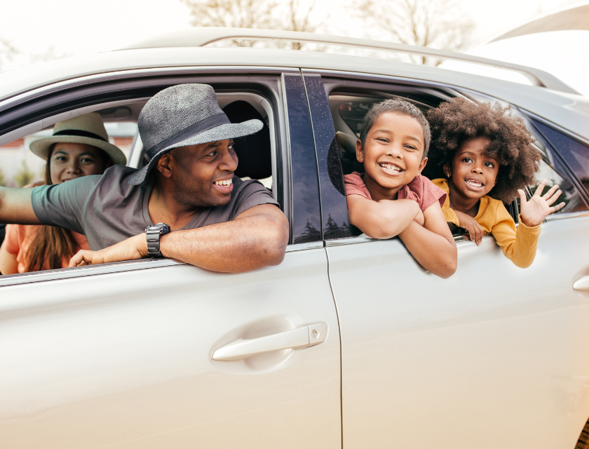 Family in a silver car. Two boys in the back and mum & dad in the front. The kids are smiling, one has an afro and the other, short hair. Parents are both wearing beach ready sun hats.