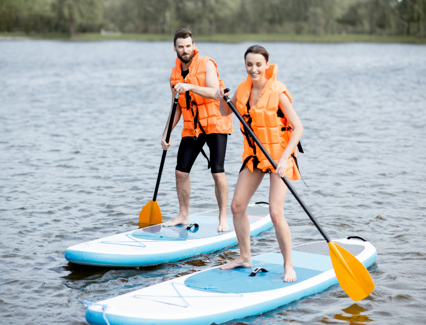 Two people (one male and one female) waterboarding on blue waterboards. They are wearing lifejackets. Out of focus trees can be seen in the background.