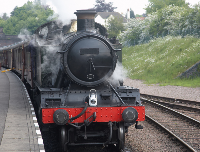 Visiting the light railway is a popular family day out & activity in Cleethorpes. A black steam train going along a track. Greenery and a house roof are visible in the background.