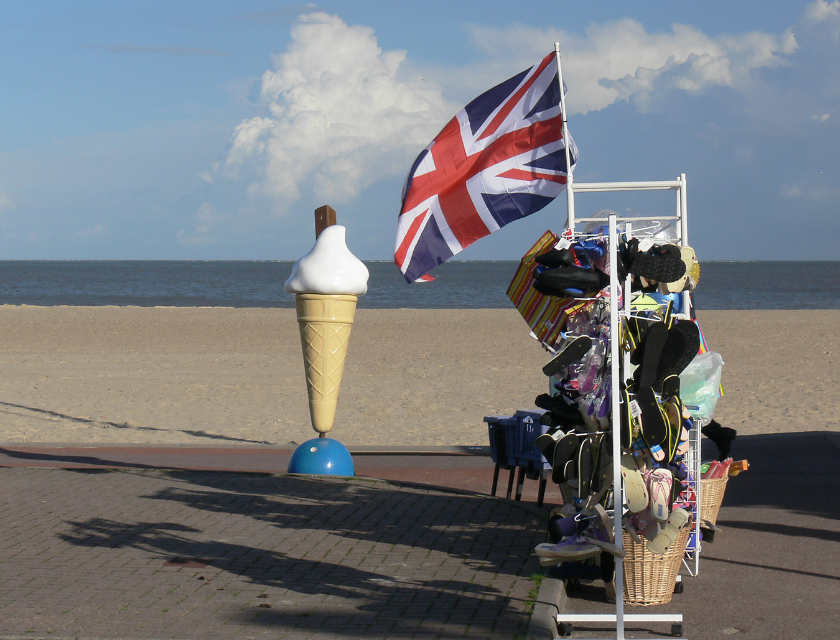 English seafront with large a plastic ice-cream, a British flag, and a stall selling hats and shoes are all visible. The shoreline, sea and sky can all be seen in the background . Several white clouds are visible in the sky.