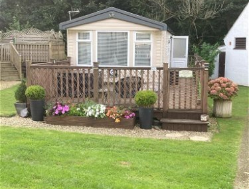 Cozy static caravan at Hele Valley Holiday Park, near Woolacombe Beach. The caravan features a a wooden deck with potted plants and flowers. There is a grassy area in front of the deck.