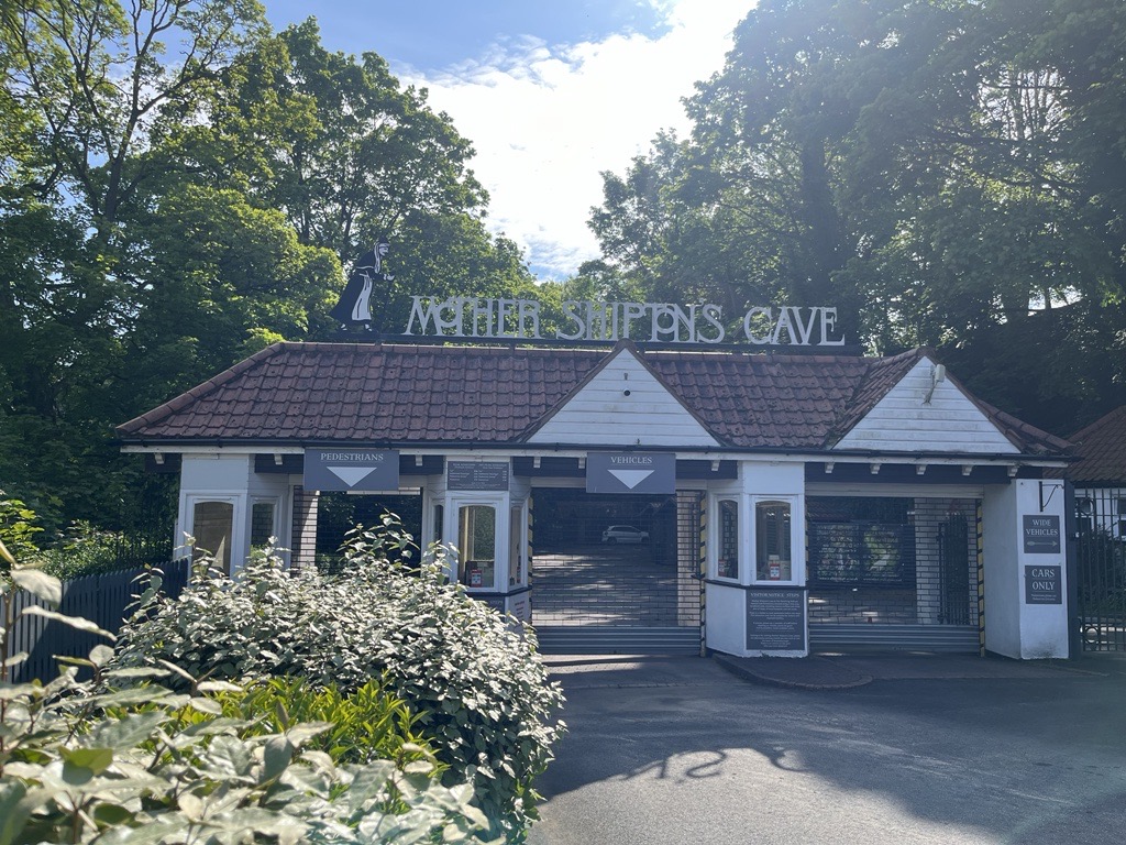 External shot of Mother Shipton's Cove in North Yorkshire. The building is backed by tall trees in-front of a blue sky.