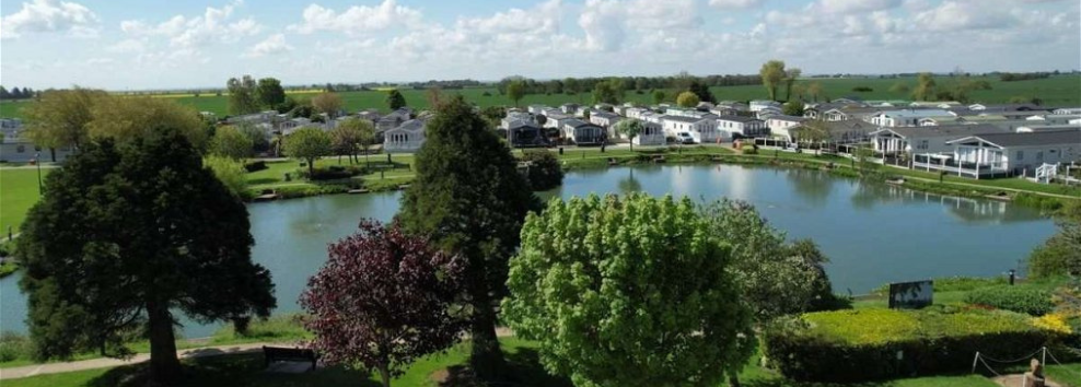 Aerial shot of a caravan park with a blue sky with white clouds throughout. Caravans are visible in the background and tall trees in the foregraound. There is a lake in between the two.