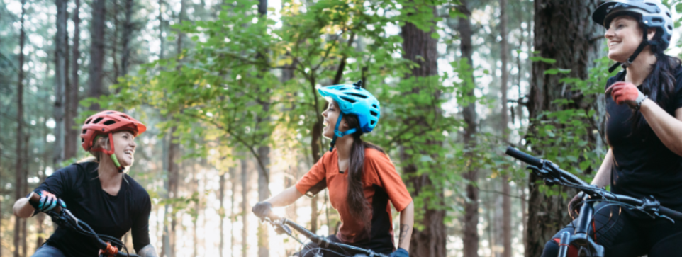 Three cyclists smiling and chatting in a forest, wearing helmets.