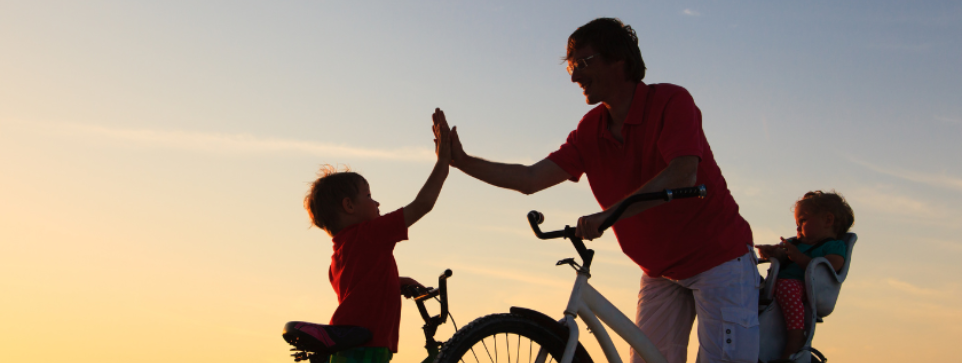 Parent and children on bikes, high-fiving at sunset with a baby in a bike seat.