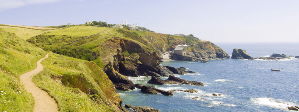 A coastal path winding along green cliffs, with waves crashing against rocky outcrops below. A quaint building sits on the edge of the cliffs, overlooking the vast blue ocean.