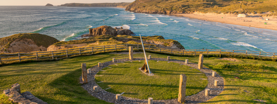 A stone sundial surrounded by standing stones on a grassy hill, with a sweeping view of the coastline and beach in the background. The scene captures a peaceful, sunny day near the sea.