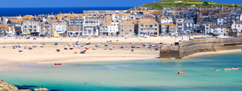 A picturesque seaside village with rows of houses and buildings featuring grey roofs and white walls. The sandy beach is dotted with boats and people enjoying the sunny day, with clear turquoise waters in the foreground.