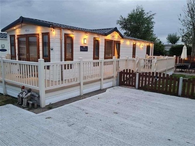 The outside of a static caravan in Withernsea at Patrington Leisure Park. The caravan is white with lights on the underside of the room creating a homely and inviting feel. There is a large decking around the caravan and a space to park in the foreground.