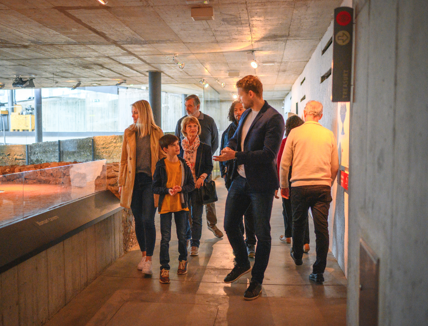 Group of visitors, including adults and children, exploring an indoor museum exhibit. The modern concrete interior contrasts with ancient ruins displayed behind glass. A guide is engaging with the group, pointing out details as they walk through the exhibit. The atmosphere is educational and interactive.