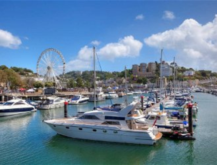 A vibrant marina in Torquay with luxurious yachts docked in the harbor, a large ferris wheel visible in the background, and clear blue skies above. The image showcases the picturesque waterfront, a popular spot for leisurely walks in Torquay.