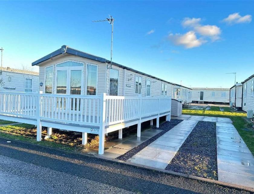 Exterior shot of a white static caravan with wrap around decking in North Wales, providing comfortable stays for visitors exploring the region's waterfall walks. There are several clouds in the bright blue sky above.