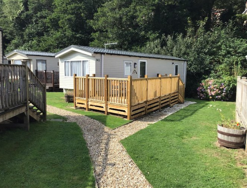 A cosy static holiday caravan with a wooden deck, surrounded by a well-kept garden and pebble path. Nestled among greenery.