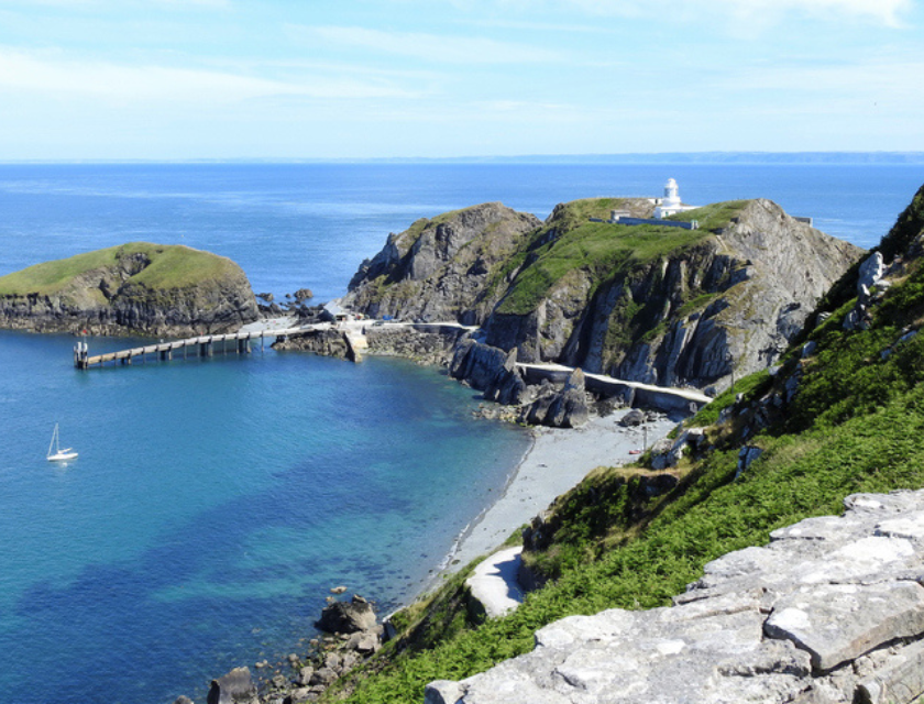 The South lighthouse on the rocky cliffs of Lundy Island. There is a white yacht moored in the harbour in the clear blue sea