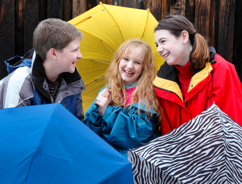 Three teenagers holding different coloured umbrellas in front of a wooden fence.