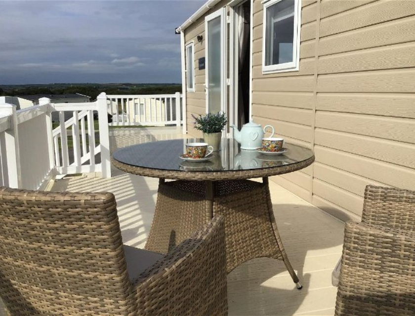 Decking of a static caravan at a caravan park, offering the perfect spot to relax after a day of walks in Boscastle, surrounded by countryside views. The exterior of the caravan is cream and the sky i visible in the background.