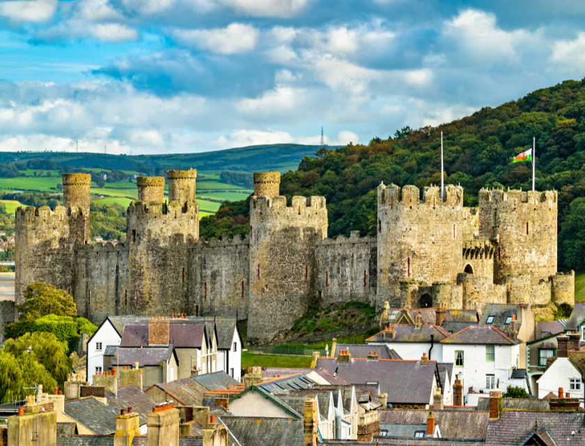 Stone castle with multiple round towers. The castle is surrounded by green hills and overlooks a village with traditional houses. The Welsh flag is flying from the castle walls.