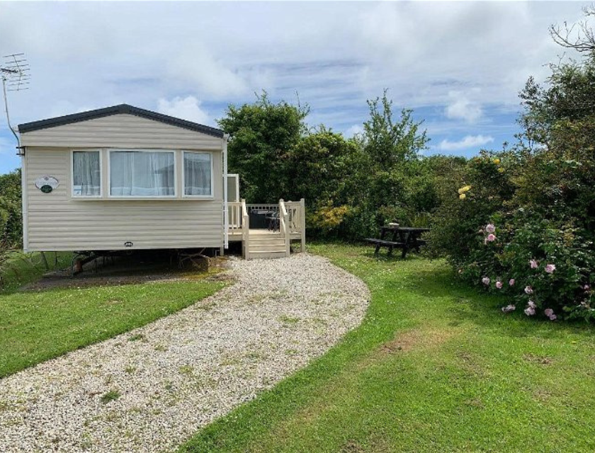 Taken at Lizard Point Holiday Park, a perfect base for dog-friendly activities in Cornwall. A cream-coloured static caravan with a small deck, surrounded by green grass and bushes, with a gravel path leading to the entrance.